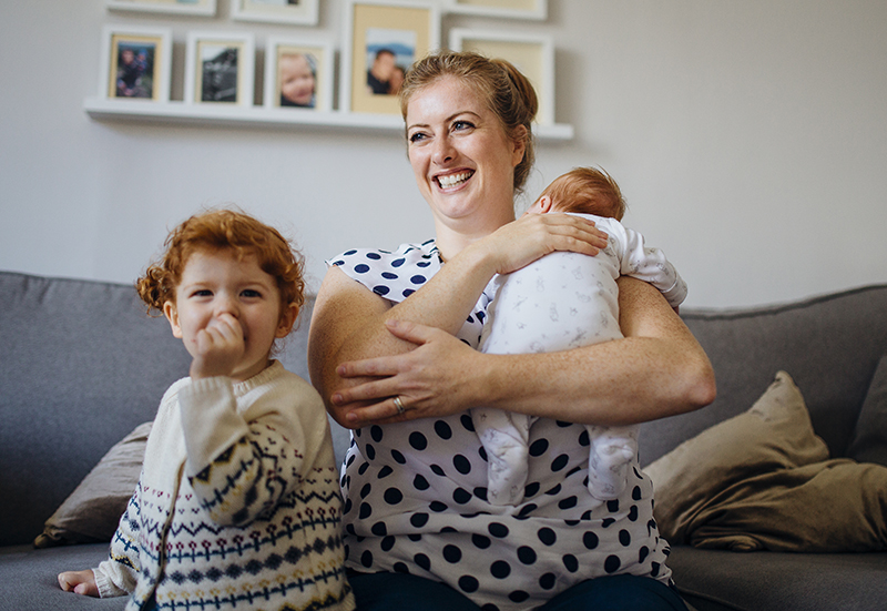 A parent sitting next to her child smiling, while holding a baby.