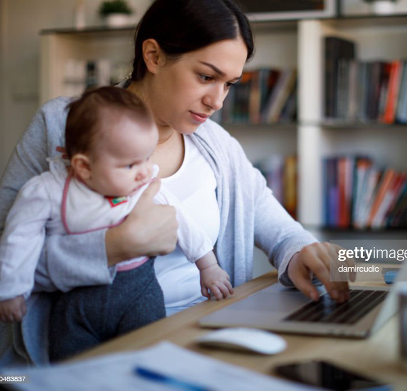 A mother working on a laptop at home whilst holding their baby