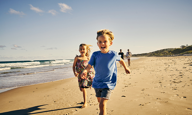 Shot of two little children running at the beach with their parents in the background Shot of two little children running at the beach with their parents in the background