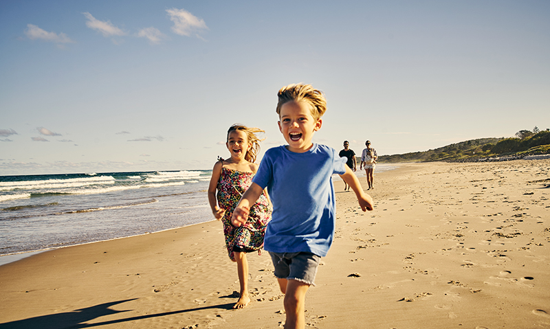 Shot of two little children running at the beach with their parents in the background