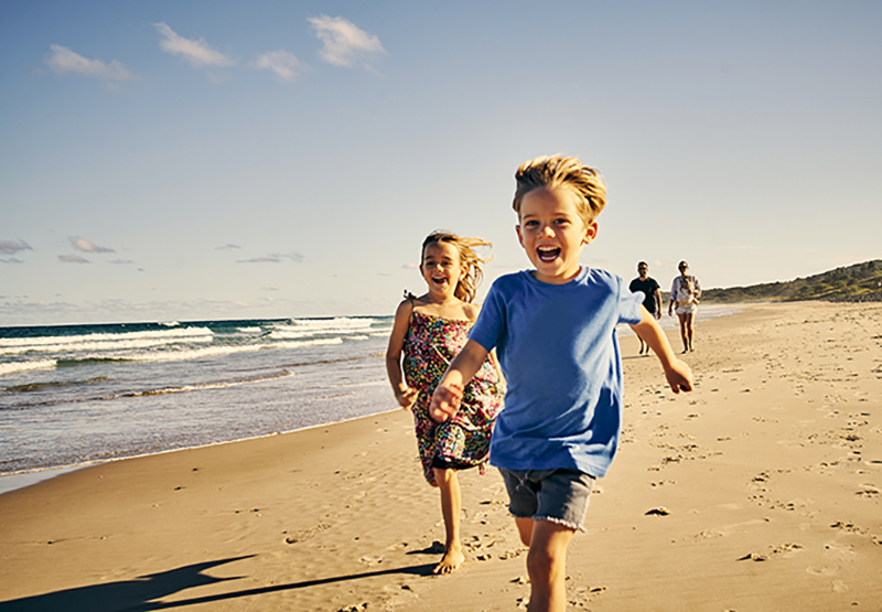 Shot of two little children running at the beach with their parents in the background