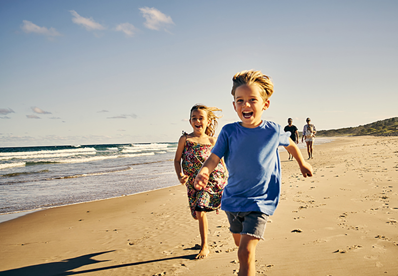 Shot of two little children running at the beach with their parents in the background Shot of two little children running at the beach with their parents in the background