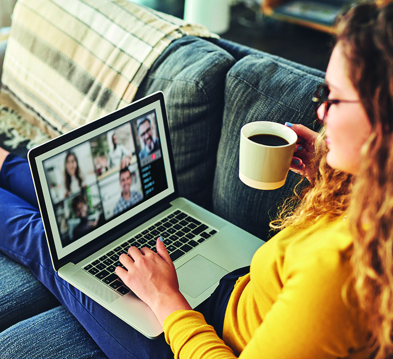 A woman attending an online meeting on a laptop whilst having coffee on the sofa at home