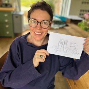 Actress Olivia Colman sat in her kitchen holding her doodle of wellies in a puddle and a smiley face.