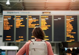Woman with her back to the camera looking at travel information on boards at the airport. She has brown hair, a pink jumper and a grey backpack on