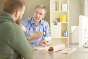 gp explains something to his male patient whilst writing a prescription
