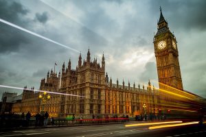 Big Ben and the Houses of Parliament in London, England. The building is lit up and set against a dramatic sky with cars and busses zooming by on Waterloo Bridge.