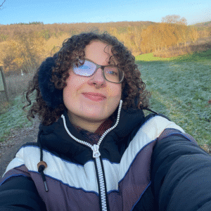 Emily a white woman poses for selfie in the outdoors she has curly brown hair and glassed and is wearing black earmuffs and a black and white zipped coat