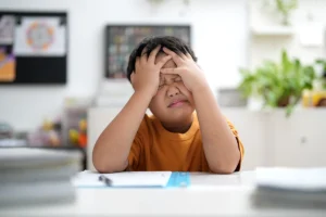 Boy in an orange shirt squishing his face in his hands, distracted and struggling with his school work