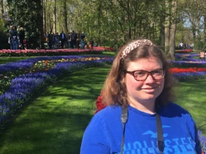 Young woman (claudia), who has autism and epilepsy, in a blue t-shirt and a hair band posing in front of a flower garden.