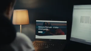 Over a person's shoulder, a screen can be seen with the Epilepsy Action website and the new report title on it. A table lamp to the left of it and the corner of a separate screen to the right.