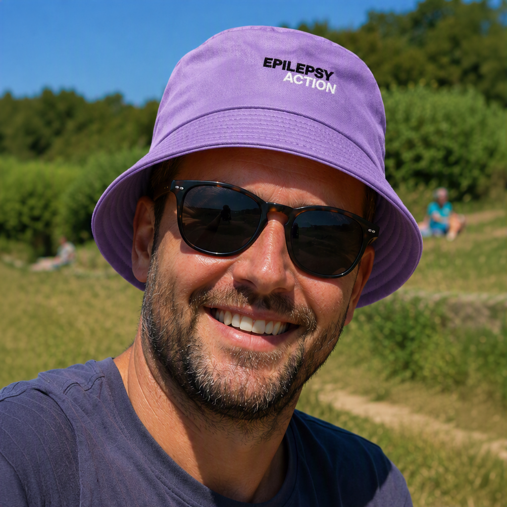 Epilepsy Action Purple Bucket Hat 1 A man is outdoors, he is wearing shades. He is also wearing a lavendar bucket hat with a black and white epilepsy action logo on the front.