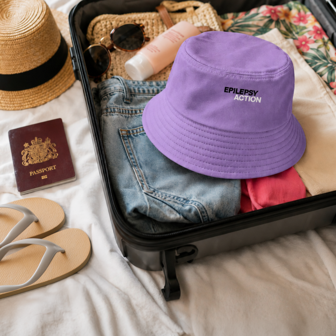 Epilepsy Action Purple Bucket Hat 2 The image shows a flat lay of a suitcase being packed on a bed. The suitcase contains holiday clothes. Alongside the suitcase are flipflops and a passport. A lavender epilepsy action hat sits on the top of the open case.