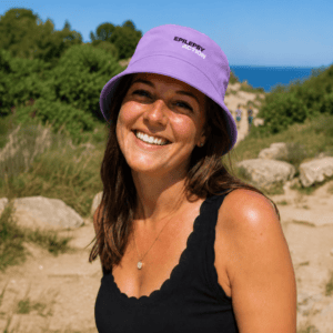 A young woman with brown hair smiles at the camera, she is wearing a lavender hat with the epilepsy action logo on the front, she is standing on a rocky beach.