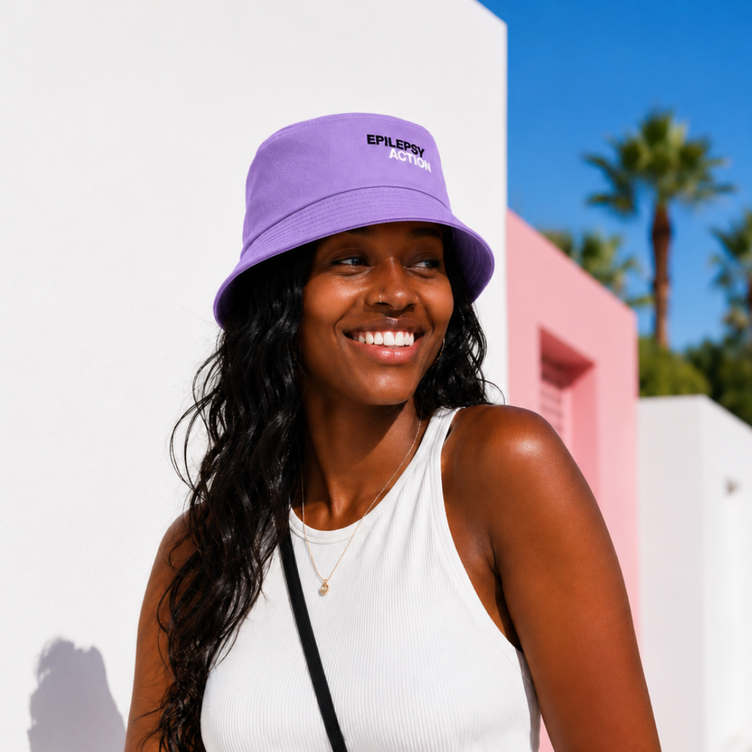 Epilepsy Action Purple Bucket Hat 3 A young black woman smiles in the sunshine. She appears to be in a sunny climate outside a building. She is wearing a lavender epilepsy action hat, her hair is in braids, she is wearing a white vest top and a gold necklace.