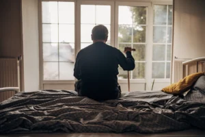 Older adults can deal with unrecognised depression. An older man, seen from behind, sat on his bed, leaning on a walking stick looking outside a window.