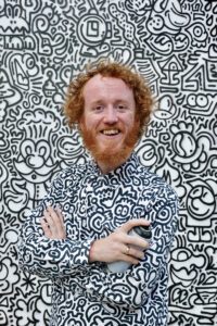 Mr doodle - a man with curly red hair smiling with a shirt covered in black and white doodles, with the same doodles on the wall behind him