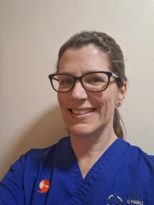 Nesta narbad. Nurse with brown hair in a pony tail and glasses smiling wearing a blue nurse uniform.