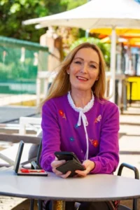 Paula mcgowan: a woman with long blonde hair sitting at a table outside and smiling. She's wearing a purple top with a white collar