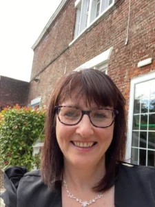 Steph morrison - woman with glasses and dark hair smiling in front of a red-brick building