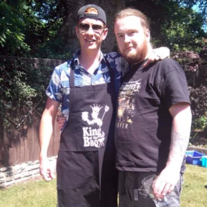 Pete and matt posing together for a photo. Pete is wearing an apron that says king of the bbq